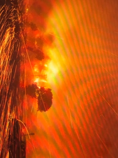 The image displays an event taking place in Kafr Sousa Municipality, Syria, dominated by intense orange and yellow illumination indicative of a large fire or explosion. On the left side of the frame, a dark, vertical structure is visible, from which numerous bright, linear streaks, consistent with sparks or incandescent debris, are actively ejecting diagonally upwards and to the right. Irregular, amorphous dark shapes are discernible within the bright orange and yellow glow, suggesting dense smoke or burning structural elements. No individuals are visible. The entire scene is engulfed in light and particulate matter, obscuring further specific environmental or structural details.