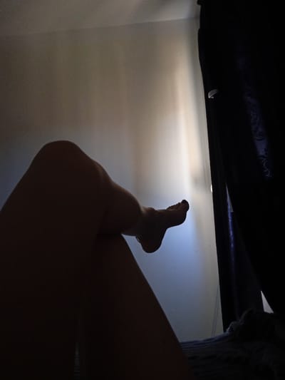 The image is of a person's legs and feet, with a shadow covering most of the body. The person is sitting in a dark room, with the legs extending out towards a wall. The wall is a light blue, and there is a shadow of a curtain in the background. The person's toes are visible at the end of the foot. The image was taken in Réghaïa, Algeria at the coordinates 36.729 latitude and 3.369 longitude.