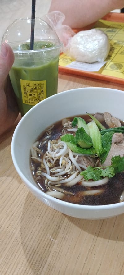 This image depicts a close-up of a meal and a beverage, likely placed on a wooden table. To the left, a clear plastic cup filled with a vibrant green beverage, possibly iced matcha, is visible. The cup has a black straw and a yellow sticker with graphic symbols and text in an unknown language. In the center and to the right, a white bowl contains a noodle soup dish. The soup has a dark broth and is topped with bean sprouts, green vegetables (likely bok choy), and pieces of meat. A sprig of cilantro is also visible as a garnish. In the background, out of focus, is a person's arm and a yellow tray with a white object wrapped in plastic, possibly food. The scene is well-lit, suggesting it was taken indoors in a casual dining setting. The presence of a person's hand indicates the meal is being consumed. The image does not provide any explicit clues to its location in the city of Khabarovsk, Russia.