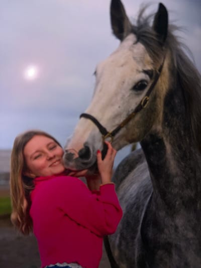 The image, rotated approximately 90 degrees clockwise, captures an intimate and joyful moment between a young person and a dappled grey horse in Estaimpuis, Belgium. A girl with light brown hair, dressed in a vibrant pink long-sleeved top and patterned blue and white clothing, is shown with her eyes closed and a wide smile, indicating deep contentment and affection. Her right hand is gently placed near the horse's muzzle as the horse nuzzles her face.

The horse has a striking appearance with a predominantly white face featuring dark grey markings around its eyes and ears, and a darker, dappled grey body. It wears a dark leather bridle. The background consists of a soft, light-colored, overcast sky, with a diffused bright spot suggesting the sun's presence, possibly indicating late afternoon or early morning light. The scene radiates warmth and a strong bond between the girl and the equine. No text is visible in the image.