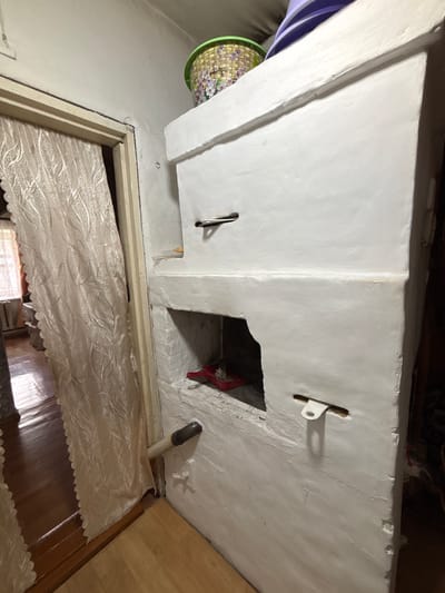 The image depicts the interior of a room, likely a kitchen or living area, in a dwelling in Ilpanur, Russia. The primary focus is a large, white, traditional Russian stove (pech) dominating the right side of the frame. The stove is constructed of plaster and appears to be functional, with visible openings and metal hardware, including what looks like a damper or vent. There's a small red shovel with a brush inside one of the stove's openings, suggesting recent use.
To the left of the stove, a doorway is visible, partially obscured by a light-colored, textured curtain with a floral pattern. Through the doorway, a glimpse of another room is offered, revealing a wooden floor and what appears to be a window with a sheer curtain. The walls in the immediate vicinity of the stove and doorway are also plastered and painted white.
On top of the stove, a woven basket with a colorful floral design is placed, and partially visible behind it are stacked purple items, possibly plates or lids. A section of the wall above the stove has a visible crack or repair. A white pipe emerges from the wall near the base of the stove and extends horizontally. The floor is covered with light-colored wooden planks. The overall impression is of a rustic, traditional living space.