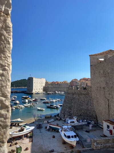 This is a high-angle, eye-level, full shot of a harbor and city walls in Dubrovnik, Croatia. In the foreground, a stone wall is partially visible on the left, with its rough texture and beige color contrasting with the bright blue sky. To the right, a section of the city's ancient stone wall dominates the frame, its weathered blocks a testament to its age.
Below the walls, the harbor is filled with numerous boats of various sizes, from small dinghies to larger yachts, many of them moored or resting on stands on a concrete slipway. The water is a clear, inviting blue.
In the middle ground, a prominent square stone tower, part of Dubrovnik's fortifications, stands guard over the harbor. Beyond it, the red-tiled roofs of the city's buildings stretch across the landscape, indicative of its historic architecture. A small island with green, treed hills rises in the background, further enhancing the picturesque scenery. The sky is a vast expanse of cloudless blue, with a few distant birds barely visible. The overall scene conveys a sense of history, tranquility, and the charm of a coastal city.
