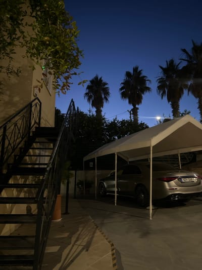 A silver Mercedes-Benz S-Class sedan is parked under a white canopy in front of a building with an exterior staircase. The scene is set at dusk or night, with a deep blue sky visible above several silhouetted palm trees. The car's license plate, "NZ Y 248," is partially visible. The staircase is made of dark metal and leads up to a building with a pale yellow facade. The ground is paved with what appears to be stone tiles. The lighting suggests artificial illumination from the building and possibly streetlights. The location is identified as Peyia, Cyprus. There are no people visible in the image.