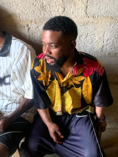 The image depicts two males seated side-by-side against a rough, light-colored concrete or plastered wall, likely within an indoor or sheltered outdoor setting in Okene, Nigeria. The prominent subject, positioned centrally, is a male with dark skin, short black textured hair with a fade, and a full black beard and mustache. He wears a short-sleeved black collared shirt featuring a pattern of large yellow and red leaves, paired with dark blue athletic shorts or trousers that have a white stripe down the side. A blue wristband with white lettering is visible on his left wrist. His mouth is slightly open, and his gaze is directed towards the right side of the frame. To his left, a second male is partially visible, wearing a white collared shirt with thin vertical black stripes and a black collar, displaying text that appears to read "OMOE" on the chest. Both individuals are seated with their hands resting on their laps. The scene is illuminated by ambient light.