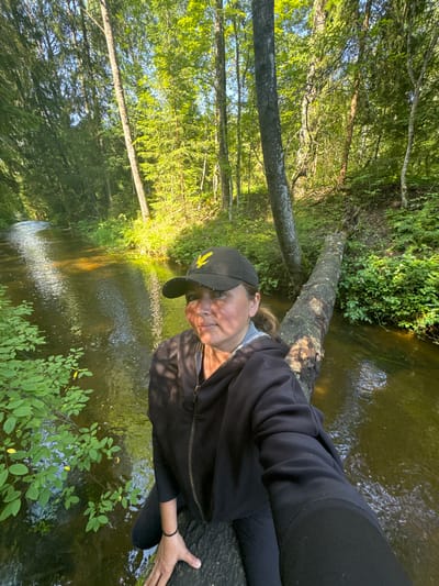 A woman is captured in a selfie on a fallen log bridging a small river in a lush, sun-dappled forest. She is wearing a black baseball cap with a yellow emblem and a black zip-up hoodie over a grey layer. Her facial expression is neutral as she looks towards the camera. The forest canopy is dense with green foliage, and sunlight filters through the leaves, illuminating the water and the forest floor. The river is shallow and clear, with a gentle current. The overall setting suggests a serene, natural environment within the city of Zelonaya Dolina, Russia.