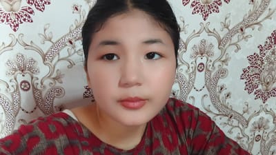 This media file is a medium close-up selfie of a young woman with dark hair pulled back, set against a backdrop of light-colored wallpaper with an ornate floral pattern in shades of red and beige. The woman is looking directly at the camera with dark eyes, a neutral expression, and subtle makeup, including lipstick. She is wearing a red and grey patterned shirt. The lighting appears to be even, suggesting an indoor setting, possibly during the daytime given the brightness. The wallpaper's intricate design hints at a traditional or decorative interior. There is no visible text in the image. The overall scene conveys a sense of personal documentation or a casual self-portrait, with the focus squarely on the subject's face.