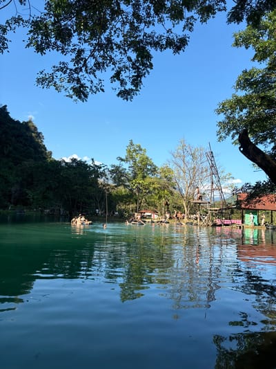 The photograph depicts a serene lake surrounded by lush green trees and a clear blue sky. A tall metal structure with people on it stands near the water, its reflection shimmering in the calm surface. Other trees and foliage frame the top and sides of the image, creating a sense of enclosure. Ripples on the water add texture to the scene. The overall impression is one of peacefulness and natural beauty.