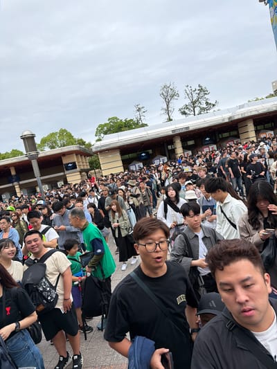 A large crowd of people, primarily East Asian individuals of varying ages from young adults to older adults, are captured in an outdoor setting, presumably queuing or waiting for an event or attraction. The image is taken from within the crowd, looking towards the front of the line which stretches into the distance alongside a multi-level, modern building.

The subjects are densely packed, standing close together. Many are casually dressed in t-shirts, jackets, and jeans, with several carrying backpacks or bags. A significant number of people are engaged with their smartphones, while others look ahead or around. In the immediate foreground, a man with curly hair appears to be resting with his eyes closed, standing alongside another man wearing glasses who looks towards the camera. One person's black t-shirt features the text "DICKIES".

The sky is bright and overcast, indicating daytime with diffused light and no harsh shadows. Bare trees with some green foliage are visible in the background and along the side of the building, suggesting a public outdoor area, possibly a park or entertainment complex. A prominent white sign with dark lettering on the building clearly reads "LOCKERS". The architecture of the building features multiple levels and railings, consistent with a large public venue.

Given the context of Osaka, Japan, and the significant crowd density with visible lockers, the scene likely depicts people waiting at a popular theme park entrance, a major event, or a tourist attraction. Despite the large number of people, the mood appears generally calm and patient.
