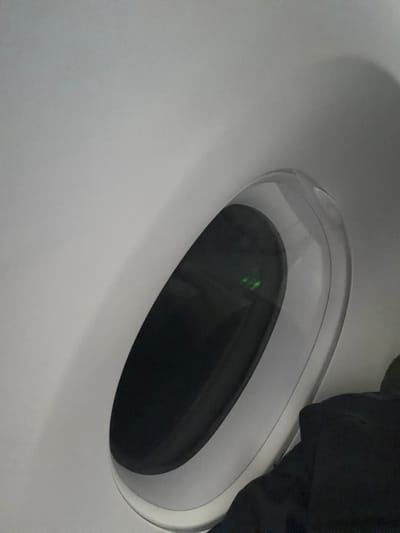 The image displays a close-up view of a recessed, oval-shaped opening set into a smooth, light-colored, possibly white or light gray, surface. This opening appears to be a window or a portal, framed by a translucent or clear rim that creates a subtle layering effect. The interior of the oval opening is dark, almost black, with several distinct, small, bright green points of light or reflections visible within it.

In the upper right portion of the image, a dark, amorphous mass, appearing to be black fabric or clothing, is visible, partially overlapping the edge of the oval opening. This could suggest the presence of a person's limb or an object like a bag resting near the opening. The overall aesthetic is modern and sleek, characteristic of contemporary design.

No specific activity is taking place, as the image is a static shot focusing on the architectural or vehicular detail. The time of day is not discernible due to the interior setting and artificial-looking light, though the darkness within the opening could suggest an exterior night scene or a deeply recessed area. No text is visible in the image.

Given the context of Addis Ababa, Ethiopia, this feature could be part of an aircraft cabin (such as a modern wide-body jet, potentially at Bole International Airport), a high-speed train, or a contemporary architectural element within a building in the city.