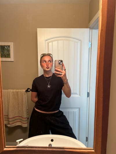 A person is taking a selfie in a bathroom. The person is wearing a black t-shirt and black pants, and has a necklace with a starfish pendant. They are holding a cell phone in their right hand. The bathroom has a white door with decorative panels, a beige wall, a towel rack with a beige and red striped towel, a white sink, and a framed picture on the wall. The photograph is being taken in Whistler, Canada.