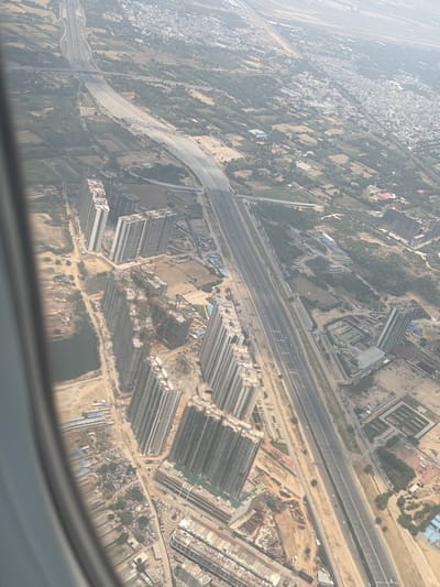 This aerial image, taken from an airplane window, showcases a sprawling urban and developing landscape in Delhi, India. The dominant feature is a multi-lane highway cutting through the scene, with visible traffic. On either side of the highway, there is a mix of existing urban areas with dense housing and significant ongoing construction of high-rise buildings, many of which appear to be under development, indicated by cranes and scaffolding. Patches of greenery and agricultural land are interspersed between the developed areas, suggesting a region undergoing rapid urbanization. A small body of water is visible in the lower left portion of the image. The overall scene depicts a dynamic urban environment with a clear contrast between established settlements, new construction projects, and undeveloped land.
