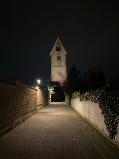 A tall stone tower with a clock face stands beside a narrow street at night. The tower is illuminated, contrasting with the dark sky. A street lamp lights the cobblestone path alongside a wall, which then meets an asphalt road bordering greenery. The perspective is angled, suggesting a view from a lower position looking upwards.