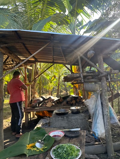 This image captures an outdoor cooking scene in the City of Carmen, Philippines, under bright sunlight filtering through dense tropical foliage. An adult male, wearing a red t-shirt and black athletic pants with white side stripes, is observed from behind, standing under a rustic shed constructed from rough-hewn wooden posts and a corrugated metal roof.

To the man's right, a stone cooking hearth with active embers supports two metal pots. Above the hearth, a substantial stack of firewood is piled on a wooden platform. Additional metal kitchenware includes an inverted pot on the firewood stack and a large grey pot suspended from a wooden beam on the right side of the shed. Two large white woven sacks are hanging from a wooden post further to the right.

In the foreground, a wooden table functions as a food preparation area. It features a large green banana leaf holding yellow fruit peelings and a metal serving spoon. Adjacent to this, a white bowl containing a red liquid, a small wooden cutting board, and a knife with a white handle are present. A white plate filled with chopped green leafy vegetables is also on the table. The background is characterized by numerous green palm fronds and banana leaves, indicating a lush, tropical environment.