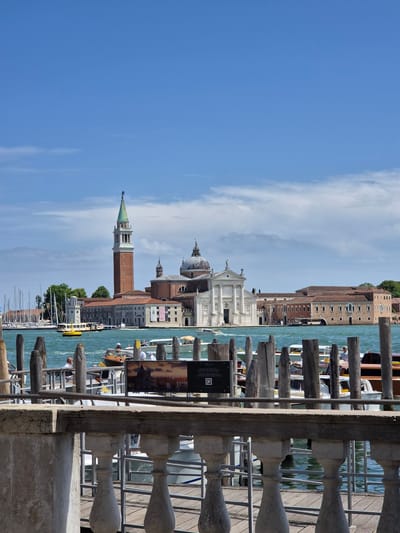 This daytime image captures a panoramic view of the Giudecca Canal in Venice, Italy, looking towards the island of San Giorgio Maggiore. The sky is clear blue with sparse, wispy white clouds scattered across the horizon.
In the mid-ground, the prominent structures of San Giorgio Maggiore dominate the scene. On the left, a tall, reddish-brown brick campanile (bell tower) with multiple arched openings rises, topped by a green conical spire and a dark, slender statue. To the right of the campanile, the white, classical facade of the Basilica di San Giorgio Maggiore is visible, featuring columns, sculpted figures in niches, and a large dome situated behind the facade. Further to the right, a series of lower, reddish-brown buildings with numerous windows, part of the monastery complex, extend along the island's waterfront. To the far left, a marina with several sailboats docked is partially visible.
The water in the canal is a choppy blue-green. A yellow vaporetto (public water bus) is visible in the mid-ground, moving from left to right and leaving a distinct wake. Various other motorboats and water taxis, some with white canopies, are scattered across the canal, several with visible passengers.
In the immediate foreground, a light-colored stone balustrade with ornate balusters runs horizontally across the bottom section of the frame. Behind the balustrade, a wooden pier or dock structure is present, featuring numerous tall, weathered, dark brown wooden mooring posts ("bricole") that extend vertically into the water. Two rectangular dark signs are mounted on the pier; the left sign shows a landscape image, text, and a QR code, while the right sign also features a landscape image. Several people are visible on the pier and in the foreground boats, including a man in a white shirt situated in a boat or on the pier in the lower-left portion of the frame.