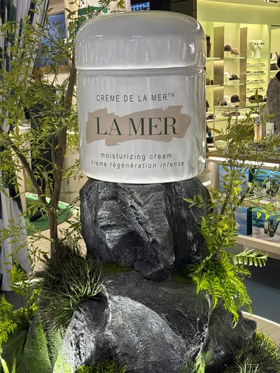 A close-up shot features a white jar of La Mer moisturizing cream prominently displayed on a rocky, mossy surface, surrounded by artificial greenery. The jar has "LA MER" in large, tan letters across its front, with "CRÈME DE LA MER™" to the left and "moisturizing cream crème régénération intense" to the right. Behind the cream jar, shelves are visible with various products, suggesting a retail or display setting. The scene is lit, creating shadows and highlights on the rocks and plants. The overall aesthetic evokes a luxurious, natural, and spa-like atmosphere, possibly within a high-end retail store or a beauty exhibition in Nong Prue Subdistrict, Thailand. The image is oriented sideways.