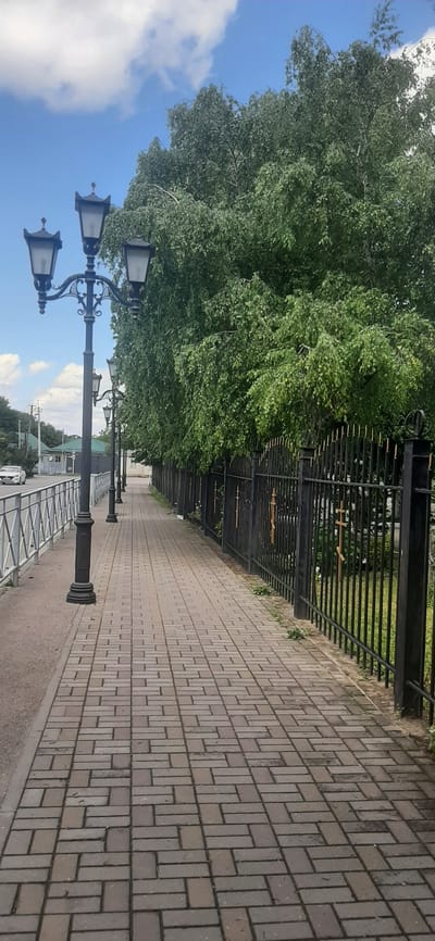 The image shows a paved sidewalk with a row of decorative lampposts on the left and a black metal fence with ornate tops and crosses on the right. A large tree with dense green foliage is visible behind the fence. The sky is blue with scattered white clouds. In the distance, a white car is driving on a road next to the sidewalk, and some buildings with green roofs are visible. The sidewalk is made of rectangular bricks laid in a herringbone pattern. The overall scene depicts an outdoor urban environment on a bright day.
