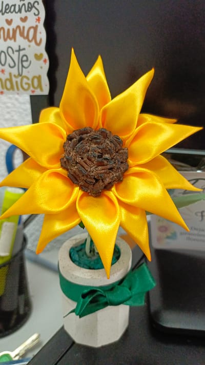 The image shows a crafted sunflower made of yellow ribbon with a brown center, placed in a small, white, geometric-shaped pot. A green ribbon is tied around the pot. The sunflower is positioned on a desk or countertop, with office supplies and a sticker visible in the background. The image was likely taken indoors. I cannot determine the location within Guayana City, Venezuela from the given information.