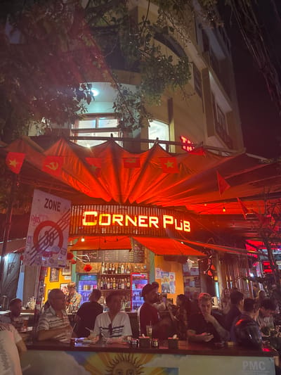 The image depicts a nighttime scene at the "Corner Pub" located in Ngũ Hành Sơn Ward, Vietnam. The pub's facade is dominated by a prominent orange neon sign displaying "CORNER PUB." An expansive red fabric canopy, illuminated from below, extends over the outdoor seating area, adorned with numerous small Vietnamese national flags (red with a yellow star) and several flags of the Communist Party of Vietnam (red with a yellow hammer and sickle).
Beneath the canopy, approximately 15-20 individuals, appearing to be adults of various ages and genders, are visible. Most are seated or standing around a long bar counter in the foreground, engaging in social interaction. Several men are seated at the counter with drinks. A black motorcycle helmet rests on the counter in the lower right. Behind the counter, a well-stocked bar with numerous bottles is brightly lit, and at least one staff member is present.
To the left of the pub's entrance, a large white banner with a "ZONE 38" logo is displayed. Small red lanterns are suspended near the bar. The pub occupies the ground floor of a multi-story building, with dimly lit upper floors visible above the canopy. Dark tree branches and foliage partially obscure the upper portion of the building and night sky. The overall lighting is a vibrant red-orange, emanating from the pub's signage and canopy, contrasting with the darker background.