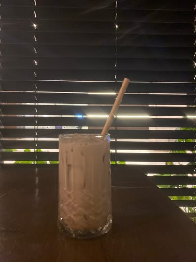 The image depicts a glass of iced coffee with a straw, resting on a dark brown table against a backdrop of black vertical blinds. Outside the blinds, hints of greenery and blurred shapes suggest an outdoor environment. The location is Rehovot, Israel, discernible by a partial view of a window displaying advertising for the Tishrei holidays in Hebrew with a phone number starting with 08, a common area code for Rehovot. No people or interactions are directly visible in the scene.
