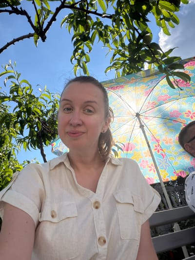The image is a close-up, selfie-style shot of a woman with light skin and brown hair pulled back in a ponytail. She is wearing a light-colored, collared, short-sleeved shirt with two pockets on the chest and light brown buttons. She is looking directly at the camera with a slight smile. Behind her, a colorful floral umbrella is partially visible, with sunlight shining through it. Green foliage from trees is also prominent in the background, with branches extending into the frame. A small, green fruit is visible on one of the branches. The sky above is clear and blue. To the right of the woman, a portion of another person, presumably a man, is visible from the shoulder up, wearing a white patterned shirt. The scene appears to be outdoors in a garden or backyard setting. The presence of a fruit-bearing tree and an umbrella suggests it is likely a warm day. The location is identified as Luka, Ukraine.