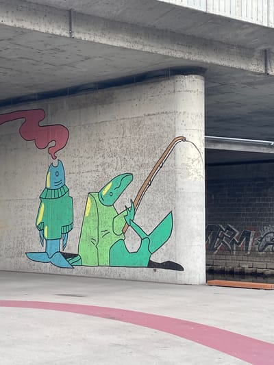 The image depicts a mural painted on a concrete wall beneath a bridge structure. The mural features two cartoonish figures. On the left, a blue, fish-like character wearing a green sweater with yellow accents and fin-like feet stands with a plume of red smoke emanating from its head. To its right, another character, also resembling a fish or amphibian with yellow accents, is depicted in a green cloak, sitting and holding a fishing rod. The fishing rod's line curves upwards.
The environment appears to be an urban outdoor space, likely a public area or park. The concrete under the bridge is rough and textured. To the right of the mural, a brick wall with graffiti is visible. The ground in front of the mural is concrete, with a pink stripe painted across it. A wooden plank or bench is placed on the ground in front of the brick wall. The lighting suggests it is daytime. This mural is located in Pilsen, Czechia, specifically on the wall of the underpass of the U Trati bridge.