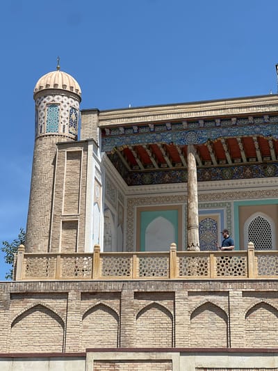 A man stands on a balcony of a building in Samarkand City, Uzbekistan, with a minaret and dome visible in the background. The architecture features intricate tilework and decorative brickwork. The man is wearing a dark shirt and a baseball cap and appears to be looking at something off-camera. The sky is clear and blue.