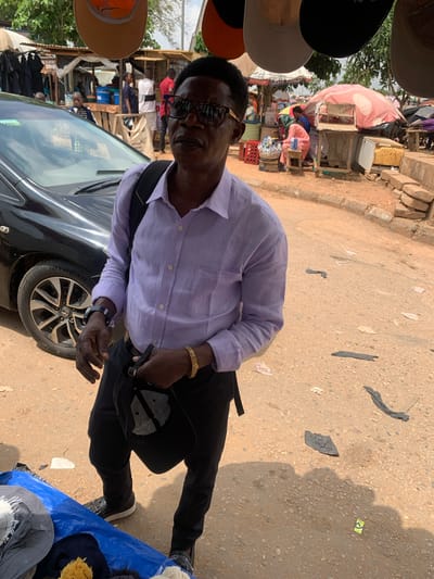 The image captures an outdoor scene, likely a market in Nyanya, Nigeria, during the daytime. The ground is a dirt and gravel surface, with scattered debris suggesting a busy, potentially dusty environment. Sunlight indicates it's a clear day.
The main subject is a man, appearing to be of African descent, standing in the foreground. He is wearing a light purple, long-sleeved button-down shirt, black trousers, and dark shoes. He also sports glasses and a gold-colored bracelet on his left wrist. He is holding a black baseball cap in his right hand, with a visible black watch on his left wrist. His expression is neutral.
Behind the man, a market stall is visible, with various items displayed. There are shelves stocked with goods, possibly clothing or accessories. In the far background, more market activity and stalls can be seen, along with trees and what appears to be a building. To the left of the man, several hats are hanging, some on display, including a light brown one. To the right, a blue tarp is spread on the ground, with more hats visible on it. A dark-colored car is partially visible in the lower-left quadrant of the image, with its wheel and part of the hood showing.
The overall impression is of a vibrant, bustling market scene, likely a typical day's trading. The man in the foreground appears to be a vendor or a shopper, engaged with the market environment.