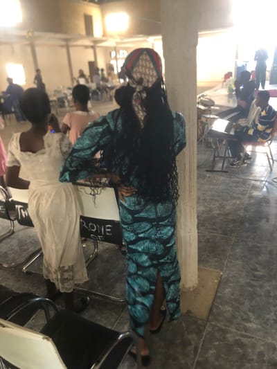 An indoor gathering is depicted within a large, light-walled hall in Samaru, Nigeria. In the immediate foreground, a woman faces away from the camera, dressed in a form-fitting teal gown with a dark, leaf-like pattern. She has long black braids and wears a patterned headscarf displaying red, green, and cream colors, possibly with a repeating "DIOR" logo. Her hands are on her hips as she leans slightly against a light-colored concrete pillar. To her left, a second woman, also facing away, wears a light-colored, possibly off-white, lace dress. A white sign on a stand between them partially displays the text "RONE OF". Several modern chairs with dark seats and chrome frames are in the foreground. In the midground on the right, two young men are seated; one plays a black electronic keyboard, while the other, wearing a striped yellow and blue shirt, observes. A partial drum set is visible behind the keyboard. Numerous blurred figures are present throughout the brightly lit background, some standing and some seated, indicating a larger congregation. The environment features light-colored walls, a tiled grey floor, and strong natural light entering from openings or windows in the background.