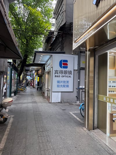 The image shows a narrow pedestrian street in Yuexiu District, China, lined with shops and buildings. The street is paved with gray stone tiles, and there are tactile paving indicators along the center. On the right side of the street is a shop with a golden facade and large glass windows displaying eyeglasses. A sign above the window reads "B&D OPTICAL" in English and Chinese characters. Further down the street, there are more shops with illuminated signs, including one with the characters "远大眼镜" which translates to "Yuanda Eyeglasses". On the left side of the street, there are awnings extending from buildings, a bus stop sign, and a few parked motorcycles and bicycles. A tree with lush green foliage grows between the buildings, casting shadows on the walkway. A woman in a red dress is walking away from the camera in the distance. The sky is overcast.