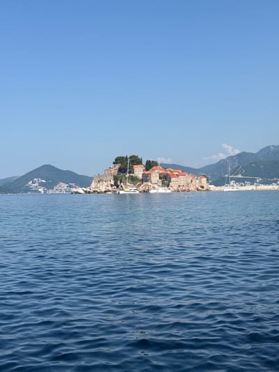 A wide-angle, eye-level shot depicts the island of Sveti Stefan, Montenegro, from the water. The island, connected to the mainland by a narrow causeway, is dotted with red-tiled buildings and lush green trees. Several white sailboats are moored in the clear blue water surrounding the island. In the background, rolling green hills and mountains rise above the coastline, with a town visible nestled among them. The sky above is a brilliant, cloudless blue. The water in the foreground is calm, with gentle ripples reflecting the sunlight.