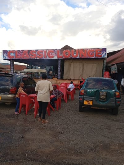 The image shows a scene outside the "Classic Lounge" in Arusha, Tanzania, located at latitude: -3.421, longitude: 36.661. The lounge has an open-air seating area with red plastic chairs and tables. A group of people are gathered, including a woman in a beige shirt and black pants standing in the foreground, a young boy in a red shirt standing to the left, a young boy in a blue shirt sitting in a chair, and a man in a blue shirt sitting at a table. A green Toyota Rav4 with Tanzanian license plates is parked to the right.  The background shows additional buildings, a few people, and a partly cloudy sky.