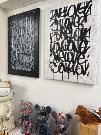 Two pieces of graffiti art hang on a white wall. The piece on the left is on a black canvas with gray and white spray paint. The piece on the right is on a white canvas with black spray paint. Both pieces feature repetitive text in a stylized script. In front of the canvases, on a white shelf, are several decorative bear figurines and a stuffed dog. The bear figurines are of various sizes and colors, with some appearing to be painted with abstract designs and others covered in graffiti. The stuffed dog is brown and is positioned on a wooden block. The scene appears to be an indoor display, possibly in a gallery or shop, given the presentation of the artwork and figurines. The overall environment suggests a contemporary art setting.