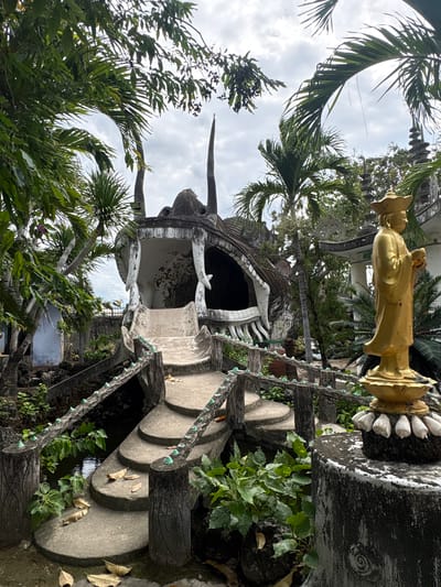 The image displays an elaborate outdoor scene, likely a cultural or religious site in Cam Ranh, Vietnam, under an overcast daytime sky. The primary subject is a large, intricately sculpted concrete structure resembling the head of a mythological creature, possibly a dragon, with its mouth agape, forming an archway or entrance. This imposing grey structure features prominent white teeth and horn-like protrusions.
Adjacent to this, on the right, stands a golden statue of a human-like figure, possibly a deity or historical persona, dressed in traditional attire and wearing a hat. The statue is elevated on a decorative pedestal which bears some faint, stylized characters that are not clearly legible.
Further to the right, a series of curved concrete steps ascend, flanked by railings that appear to be fashioned from dark, textured material with embedded small, light-colored decorative elements, resembling chains. The entire scene is richly enveloped by lush green vegetation, including numerous tall palm trees whose fronds are visible against the cloudy sky, and various bushes and smaller plants growing around the structures and steps. No human activity is depicted. The cloudy sky suggests diffused daylight rather than direct sun.