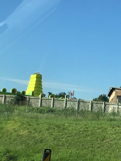 A yellow water slide tower stands beside a wooden fence line bordering green grass under a clear blue sky. Behind the fence is a small building and a playground structure. A section of the photograph is blurry, making it difficult to discern specific details. The overall scene suggests an amusement park or recreational area.
