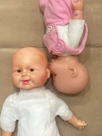 Two baby dolls are positioned on a beige, textured surface, which appears to be a sofa or carpet. One doll, in the foreground and oriented upright, has a bald, light-skinned head with brown eyes, a slightly open mouth showing teeth and tongue, and rosy cheeks. It is wearing a white, textured garment with puffed sleeves. There are some faint red marks on its forehead. The second doll, in the background and upside down, is wearing a pink hooded garment with white trim. Its head is turned away from the viewer, with only its ear and the back of its head visible. Purple marks, possibly from marker, are visible on its arm. The overall scene is brightly lit, suggesting an indoor setting. There is no information available to determine the location of this scene in the city of Dolgoprudny, Russia.