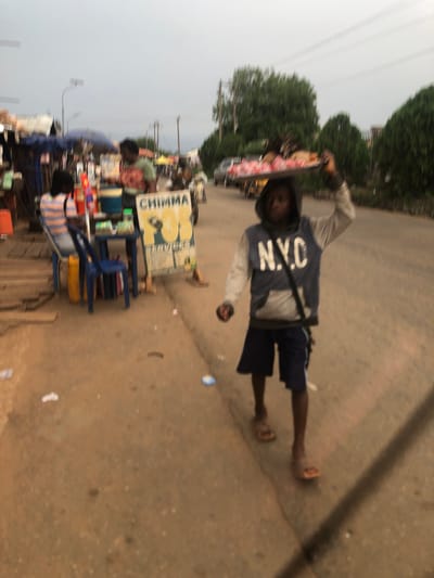 The image shows a street scene in Nsugbe, Nigeria. A young man wearing a grey and blue "N.Y.C" hoodie, dark shorts, and sandals is walking on the road, carrying a tray with items on his head. He has a strap across his chest. To the left, there is a makeshift roadside stall with a sign that reads "CHIMMA" and displays items for sale, including drinks and snacks. People are standing around the stall. The road is paved and appears somewhat worn. Trees and power lines are visible in the background, and the sky is overcast.