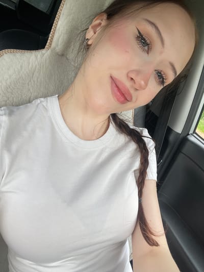 A young woman with fair skin and brown hair braided into a single plait is shown from the chest up, inside a vehicle. She is wearing a white t-shirt. She has applied makeup, including winged eyeliner and pink blush on her cheeks, and is wearing hoop earrings. Her head is tilted, and she is looking towards the camera. The background consists of a car's interior, including a cream-colored fuzzy car seat cover and a black car door. The image appears to have been taken during the daytime, as suggested by the lighting. There is no visible indication of the location being Gavrilovka, Russia.