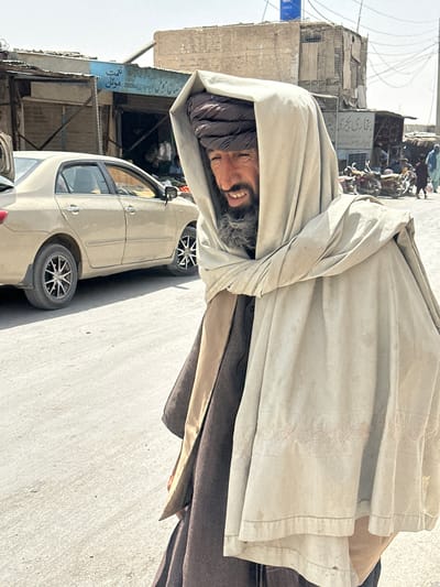 In the city of Chaman, Pakistan, a man with a dark beard and a turban is seen in the foreground, wearing a light-colored shawl and a brown garment. He is facing away from the viewer, looking to his right with a slight smile. Behind him, a beige sedan is parked on a dusty road. Shops line the street, with their shutters down. In the background, buildings made of concrete and corrugated metal are visible, along with a tangle of electrical wires. Several motorcycles are parked in front of one of the shops, and a few other people can be seen in the distance. The scene appears to be a bustling street in a busy market area.