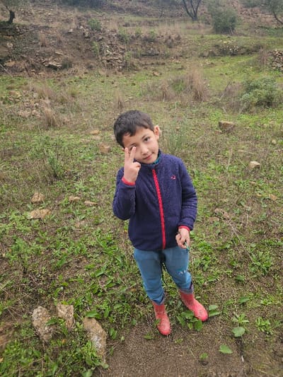 A young child stands outdoors in the city of Moqrisset, Morocco, looking directly at the camera. The child wears a dark blue fleece jacket with a red zipper, light blue sweatpants, and red rubber boots. The child's right hand forms a peace sign (V-sign) held near the right eye, while the left hand holds a small green object. The ground beneath the child consists of dark soil, transitioning to sparse green vegetation and scattered rocks. The background features an upward-sloping terrain with more exposed rocky soil, dry brown vegetation, and a few distant, sparsely foliated trees or shrubs under what appears to be an overcast sky.