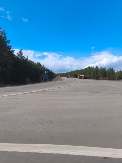 The image captures a bright, daytime scene of a paved road winding through a forested landscape in Gol'yany, Russia. The road, made of asphalt, dominates the foreground and mid-ground, showing a gentle curve towards the left. A solid white line marks the edge of the road on the right side.
Dense evergreen forests, primarily coniferous trees, line both sides of the road, stretching from the mid-ground into the background. The sky above is a clear, vibrant blue, dotted with scattered white cumulus clouds, indicating fair weather and bright sunshine, likely midday.
In the mid-ground, an orange dump truck with a flatbed is parked or stopped on the right side of the road, near a turn-off. Farther down the road on the left, a smaller, dark-colored car is also visible. Adjacent to the forest on the left side of the road, two signs are erected. The prominent one is a blue rectangular sign with partially visible white or yellow text, including characters that resemble "3C" or "АЭС" (AES) and the number "30". A smaller, circular sign is positioned above it. No people are visible in the frame. The scene portrays a tranquil, natural environment with vehicular presence, suggesting a travel route.