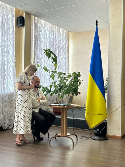 This media file captures an indoor scene featuring two individuals, a man and a woman, in what appears to be a formal or semi-formal setting, possibly a hall or meeting room, in Myronivka, Ukraine. The Ukrainian flag is prominently displayed, suggesting a patriotic or official context.
The man is seated, wearing a light-colored shirt and dark trousers. He has a beard and appears to be looking towards the woman. The woman, standing, is dressed in a white dress with black polka dots and is also facing the man, with her hands clasped. Their body language suggests an intimate or engaged conversation.
In the background, there is a plant with green leaves and a red flower, adding a touch of nature to the indoor environment. The room is decorated with white drapes that have a textured pattern. A speaker is mounted on the wall. The lighting suggests it is daytime. The overall mood appears to be calm and conversational. No visible text is present in the image.