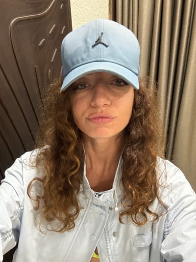 A woman with curly auburn hair is wearing a light blue baseball cap with a Jordan logo and a light blue jacket with silver snap buttons. She is looking at the camera. The background consists of a dark brown surface with linear indentations and a beige curtain with horizontal folds. The location is Sa Pa Ward, Vietnam.
