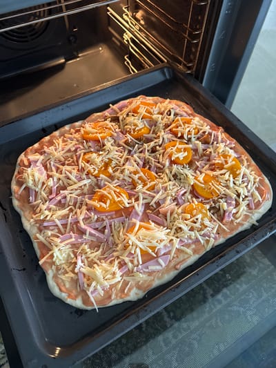 A rectangular pizza is on a baking sheet, partially inside an oven. The pizza has a tomato sauce base, topped with sliced ham, yellow bell pepper slices, and shredded cheese. The oven is dark, with oven racks visible. The baking sheet is dark gray. The lighting suggests an indoor setting, likely a kitchen. The image does not contain any elements that would allow for the determination of its location in the city of Мичуринское, Russia.