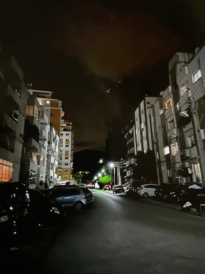The image depicts a nighttime street scene in Harmanli, Bulgaria, lined with multi-story apartment buildings on both sides. Several cars are parked along the street, and some apartment windows are illuminated. The sky is dark and cloudy, with a few faint lights visible in the upper portion of the frame, possibly indicating aircraft or distant lights. The street is paved and appears to be residential. The overall impression is that of a quiet urban environment at night.