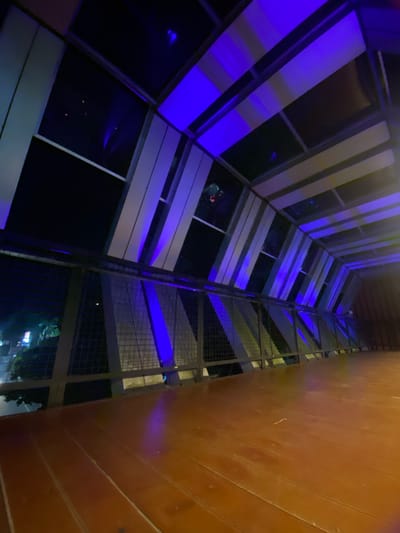 The media file captures an upward view of a modern building's exterior at night. The building features a facade with alternating panels of light-colored material and dark windows. Blue LED lights are illuminating some of the panels, creating a striking visual effect. Adjacent to this facade is a metal grate structure, possibly a walkway or part of the building's internal scaffolding, also lit by the blue lights in sections. To the right, a portion of a wooden-paneled wall is visible, its texture and color suggesting an outdoor setting or a transition to another architectural element.
The scene is set at night, as indicated by the darkness and the artificial lighting. There are no clear subjects like people or distinct objects, but reflections in the dark windows hint at the surrounding environment or interior lights. The overall mood is contemporary and somewhat dramatic due to the intense blue lighting against the dark backdrop. Location cues like "Banjarbaru, Indonesia" are provided but are not visually discernible within the image itself. No visible text is present in the media file.