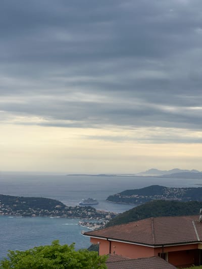The image captures a panoramic coastal landscape from a high vantage point, overlooking the French Riviera near Èze. The perspective is broad, encompassing elements from a foreground building to distant mountains and a vast expanse of sea and sky.

In the immediate foreground, part of a building with a distinctive reddish-brown tiled roof is visible on the right side. Adjacent to it, another section of a similar structure suggests a larger complex, possibly a villa or hotel. Lush green foliage from a tree frames the very edge of the right side, providing a natural element.

The midground features a prominent bay with deep blue water, characteristic of the Mediterranean. A significant landmass, heavily covered in dark green vegetation, extends into the bay, forming a peninsula. Scattered along its slopes and coastline are numerous smaller buildings, likely residential or resort properties, adding a touch of human presence to the natural beauty. A large, multi-decked cruise ship is anchored or slowly navigating in the bay, a common sight in this popular tourist region. Smaller boats can also be discerned on the water.

In the background, successive layers of landmasses, possibly mountains or distant coastlines, fade into a soft, hazy horizon, blending with the sky. The sky itself is dominated by a sweeping expanse of clouds. Closer to the horizon, the clouds appear brighter and more diffuse, suggesting the presence of indirect sunlight. Towards the top of the frame, the clouds become denser and darker, creating an overcast but not stormy atmosphere.

There are no people visible in the image, and the primary activity is the scenic display of nature and human settlement along the coast, with the cruise ship hinting at ongoing maritime travel or tourism. The light suggests it might be an overcast day, or possibly early morning or late afternoon, given the softer illumination. No visible text is present.