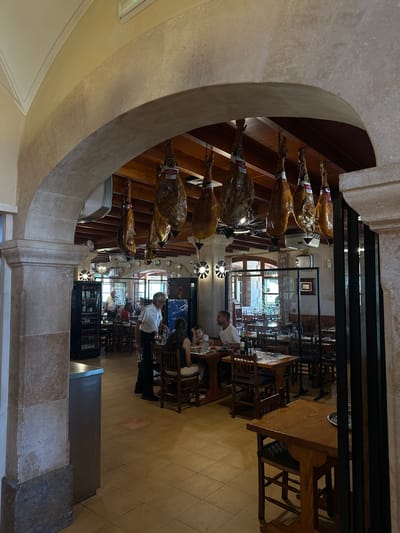 The image depicts the interior of a restaurant, likely located in Palma de Mallorca, Spain, a common misconception being Gènova which is a different region. The scene is framed by a stone archway, with several legs of cured ham hanging from the ceiling. Inside, diners are seated at wooden tables, some interacting with each other and others with staff. A waiter in a white shirt is seen serving at one table, while other patrons, including men, women, and children, are engaged in conversation or eating. The restaurant features a rustic aesthetic with wooden beams on the ceiling, tiled floors, and traditional wooden furniture. A glass-enclosed wine display is visible on the left side of the image, and ambient lighting creates a warm atmosphere. The overall impression is that of a traditional Spanish dining establishment.