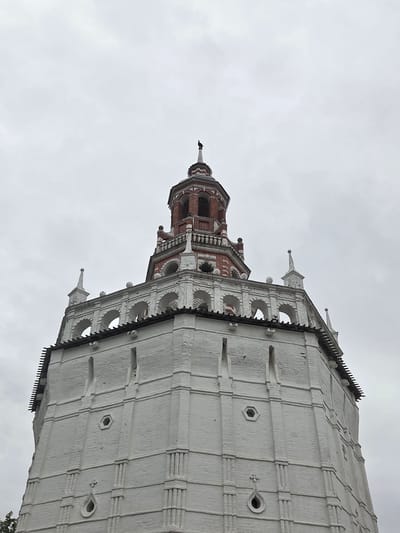 The image shows the bell tower of the Trinity Lavra of St. Sergius in Sergiyev Posad, Russia. The bell tower is a tall, white brick structure with a red-tiled roof and a spire. It is octagonal in shape and features multiple levels, each with arched windows. The top of the bell tower is adorned with decorative brickwork and several small spires. A dark bird is perched on the very top of the spire. The sky is overcast and gray. The image is taken from a low angle, looking up at the bell tower, emphasizing its height and grandeur.
