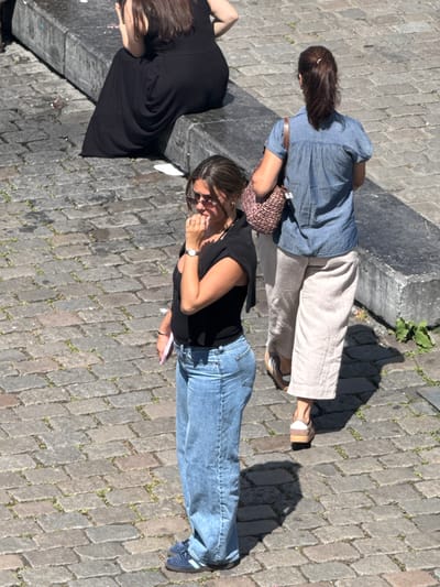 In Ghent, Belgium, a high-angle shot captures three women on a cobblestone street. In the foreground, a woman in a black shirt and blue jeans stands with her hand to her mouth, looking downwards. To her right, a woman in a blue denim shirt and tan trousers walks away from the camera, carrying a woven bag over her shoulder. In the background, a third woman in a long black dress sits on a concrete ledge, her back to the camera. The scene is lit by bright sunlight, casting distinct shadows on the cobblestones.