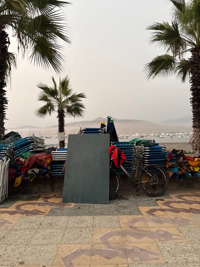 The image shows an outdoor scene likely in Ancón, Peru, as suggested by the architecture and environment. The viewpoint is tilted to the right. The sky is overcast and hazy. The foreground and background feature sections of palm trees. A row of stacked blue beach chairs and other beach-related items are stacked together, and a bicycle leans against them. A sign appears next to the stacked chairs. To the right is a patterned sidewalk. In the distant background are mountains and a faint outline of the beach.